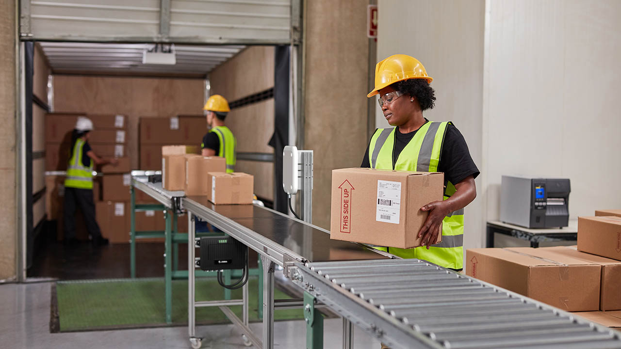 Warehouse worker in a hard hat and safety vest carrying a labeled box beside a conveyor at a loading dock